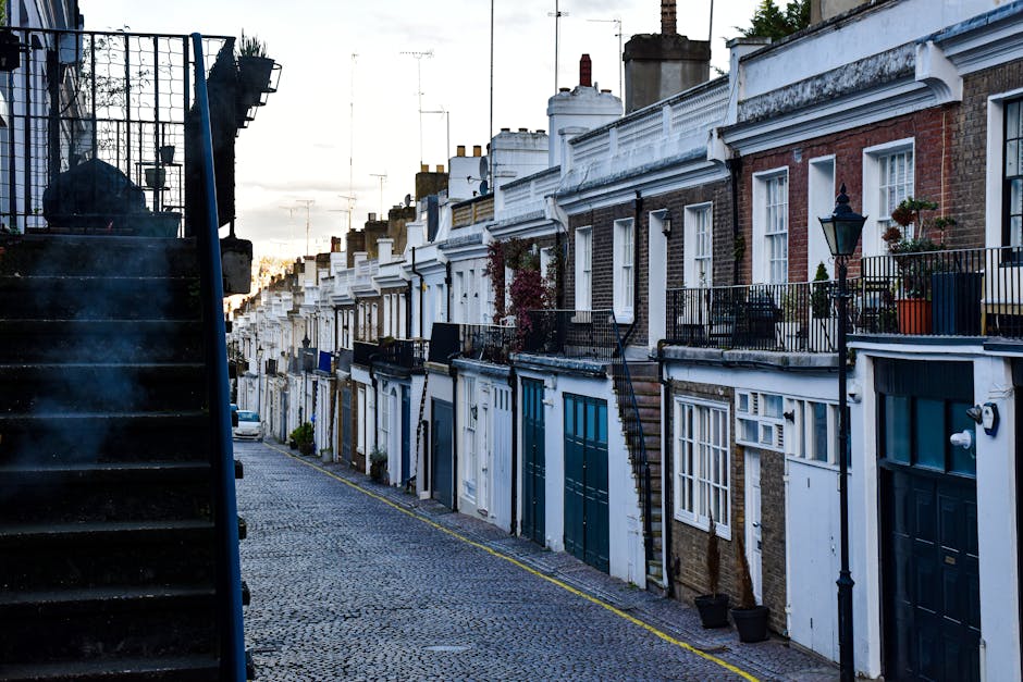 A narrow cobblestone street with a slight incline, lined with row houses featuring white and brick facades, large windows, and small front gardens with potted plants and decorative railings. Some hous