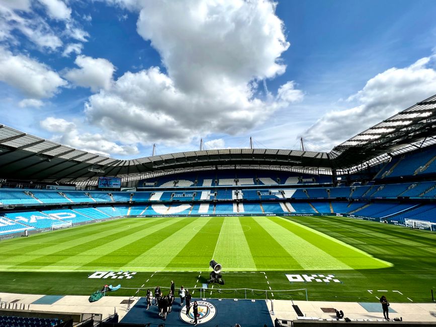 A section of Twickenham Stadium seating with rows of bright blue plastic chairs, interspersed with a few white chairs forming a patterned design. Behind the seating area, visible banners display team 